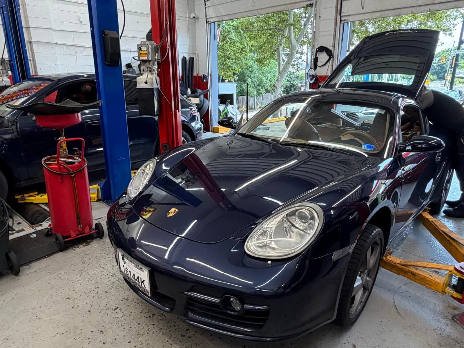 A technician working at the open rear engine lid of a dark blue Porsche Cayman in the bay, with an oil drain caddy alongside and the Langston Blvd street sign visible through the open door